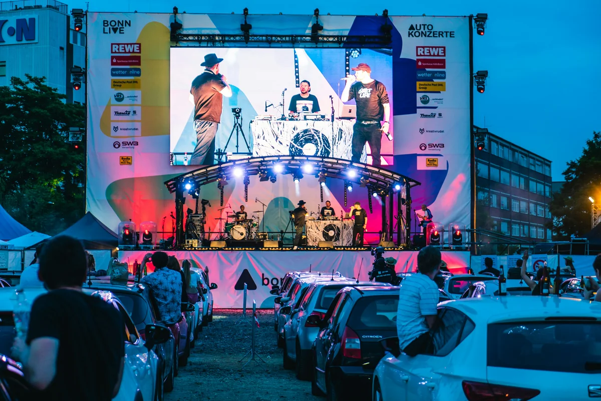 car touring, people stay in the car and see the outdoor live shows. a led video wall hanging on the stage and singer stand on the stage.