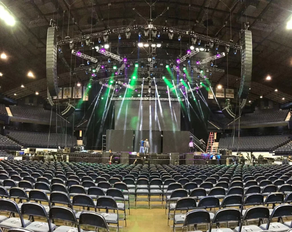 Concert stage featuring a large LED display, three side LED screens, green and red lighting effects, professional audio system, and rows of black audience chairs.