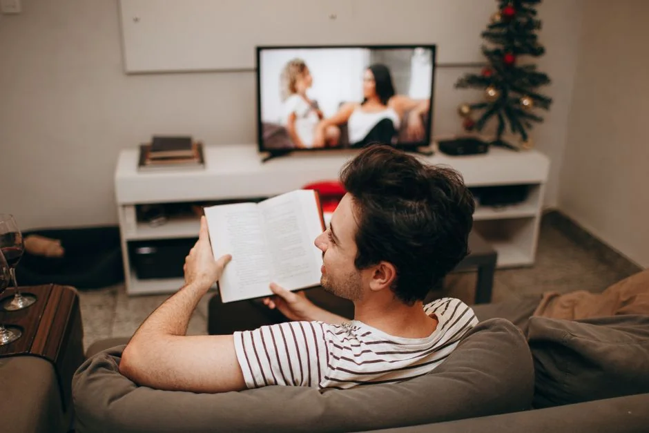 A man sits on a sofa reading a book in a cozy living room with a TV on. - home jumbotron